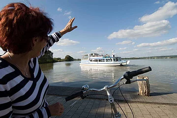 Eine Fahrradfahrerin grüßt die Passagiere der MS Altmühlsee vom Ufer aus Eine Fahrradfahrerin winkt vom Ufer aus der MS Altmühlsee zu.