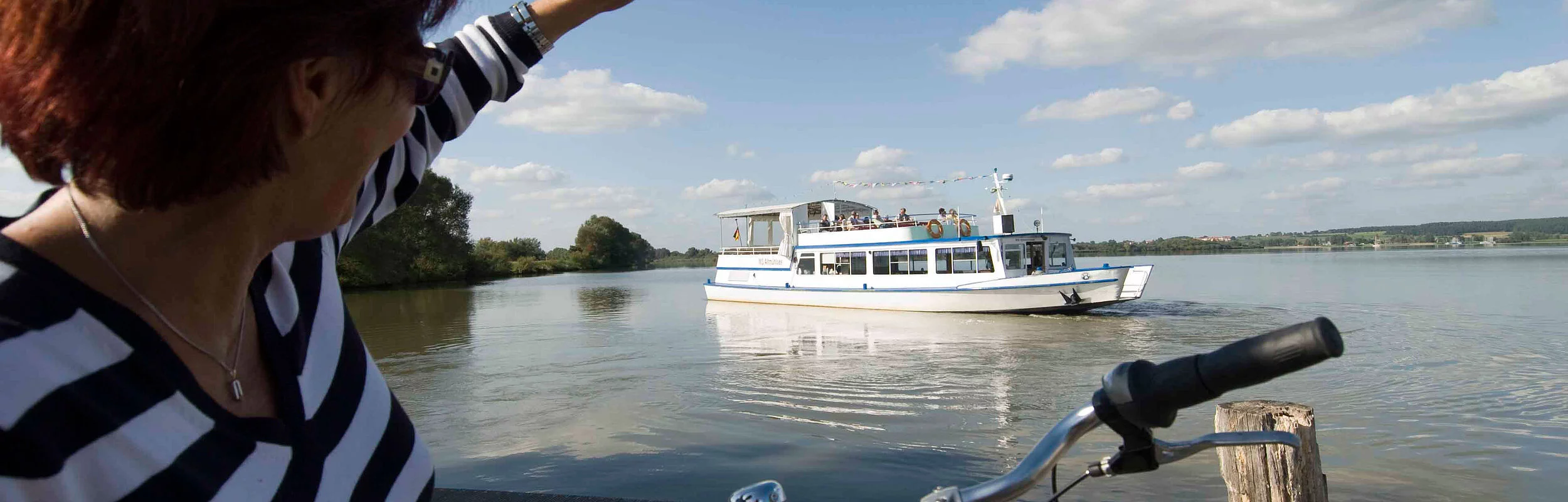 Eine Fahrradfahrerin winkt vom Ufer aus der MS Altmühlsee zu.
