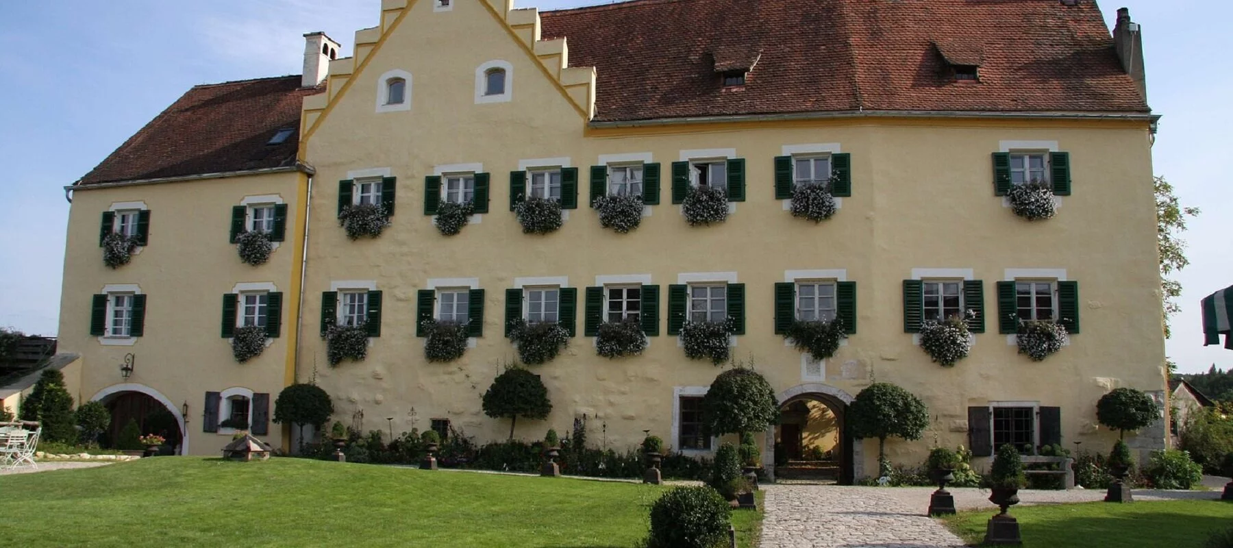 Das imposante Schloss mit seiner hellgelben Farbe und grünen Fensterläden. Davor geht ein gepflasterter Weg auf den Eingang zu. Rechts und links eingerahmt von grünem Gras.
