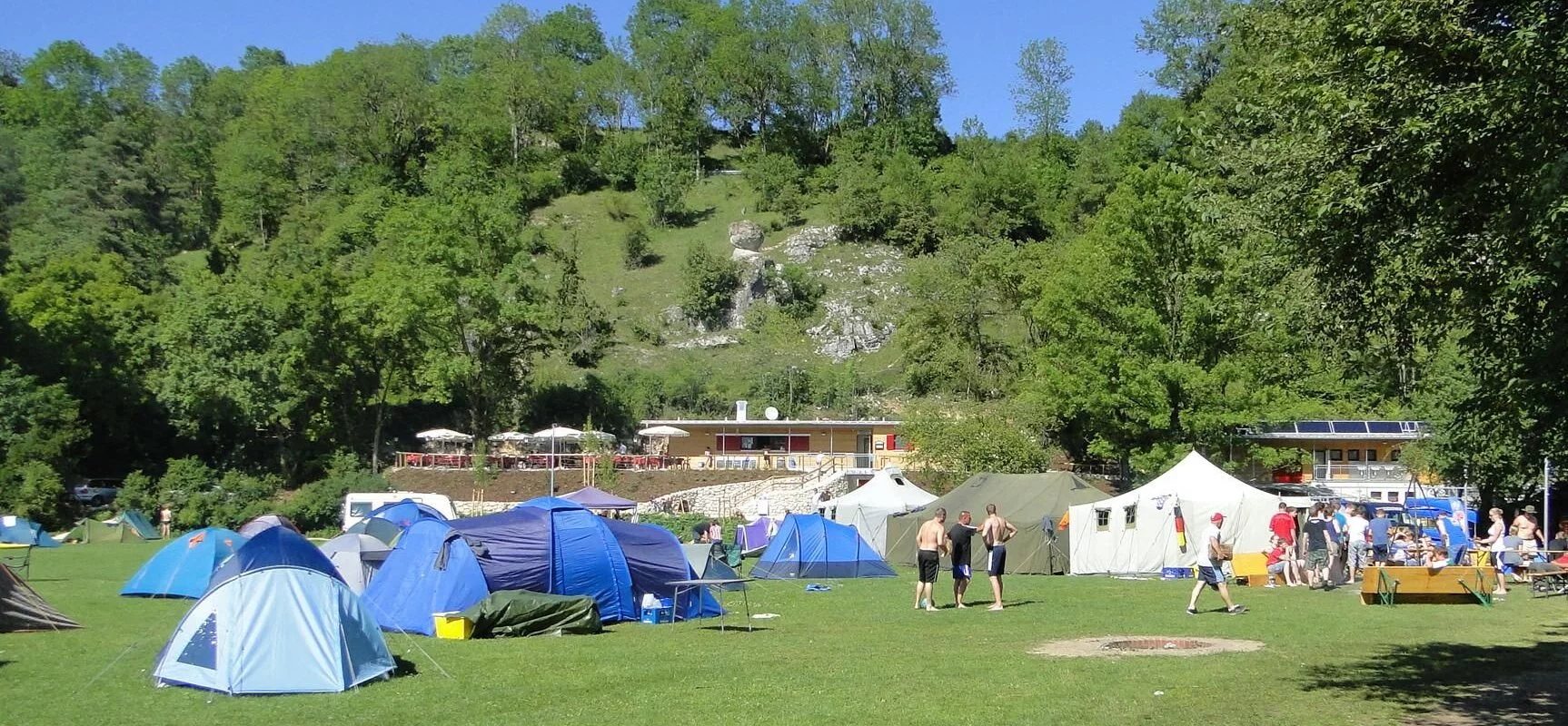 Der Zeltplatz mit vielen Zelten. Im Hintergrund die Bäume und der Himmel strahlt hellblau.