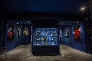 In der Mitte steht eine große Vitrine, die blau erscheint. Dahinter hängen an den Wänden rechts und links Gemälde.