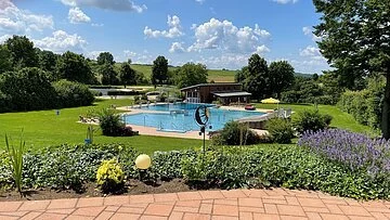 Familien- und Freizeitbad Langenaltheim Das Schwimmbecken ist umgeben von grünen Wiesen und einigen Bäumen, die Schatten spenden. Dahinter ist ein Holzhaus zusehen. Der Bereich davor ist gepflastert.