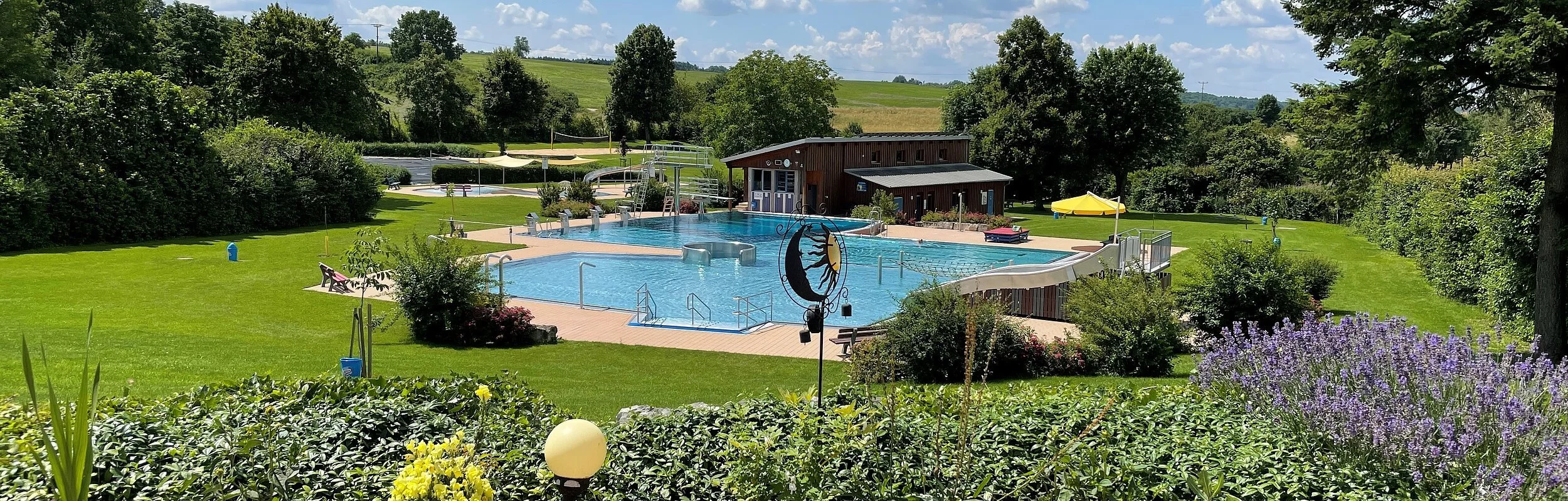 Das Schwimmbecken ist umgeben von grünen Wiesen und einigen Bäumen, die Schatten spenden. Dahinter ist ein Holzhaus zusehen. Der Bereich davor ist gepflastert.