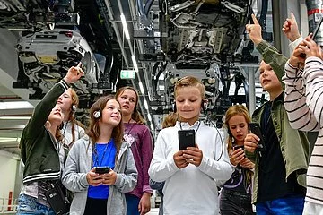 Eine Kindergruppe ausgestattet mit einem Audioguide erleben eine Werkbesichtigung bei AUDI. Die Gruppe steht unter einem Auto und betrachtet das Fahrzeug sehr interessiert.
