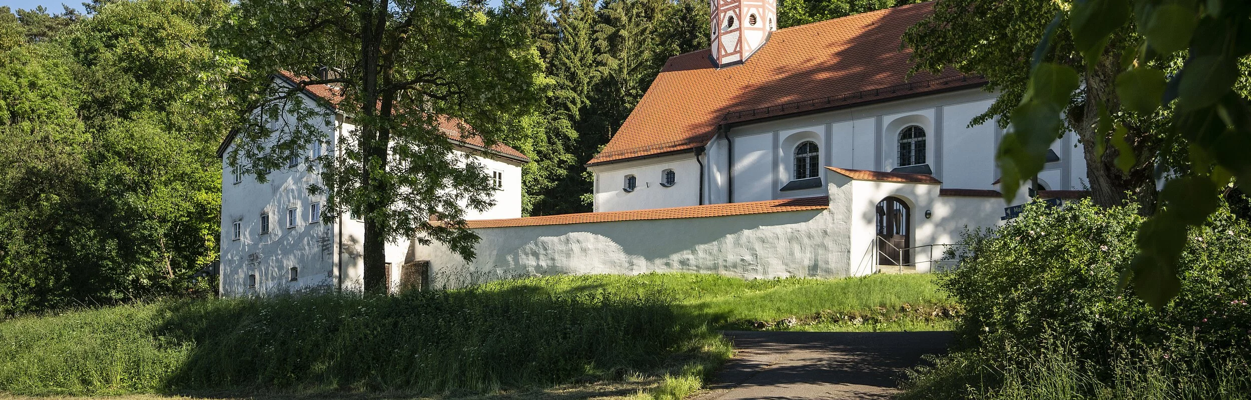 Die Wallfahrtskirche mit ihrem weißen Gemäuer und roten Dach ist umgeben von grünen Wäldern und Gras. Der Himmel scheint strahlend blau.