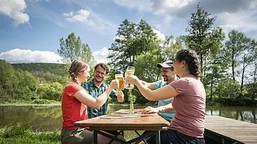 Zwei Pärchen sitzen an einer Biertischgarnitur und prosten sich mit gefüllten Biergläsern zu. Im Hintergrund ist die Altmühl und grüne Bäume zu sehen.