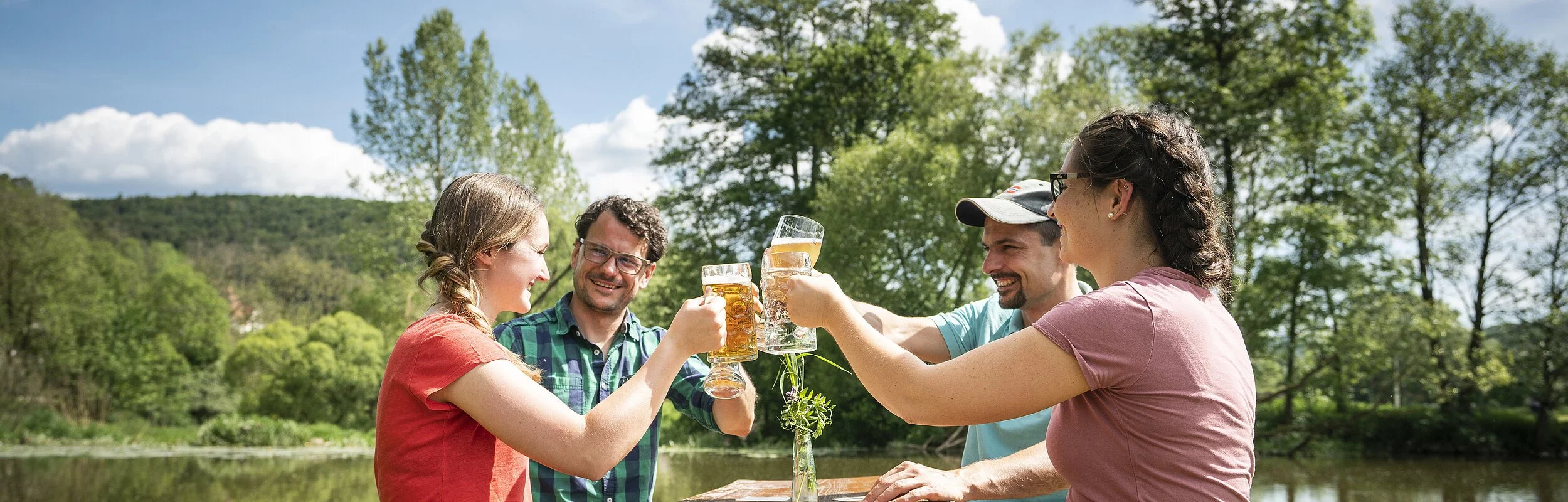 Zwei Pärchen sitzen an einer Biertischgarnitur und prosten sich mit gefüllten Biergläsern zu. Im Hintergrund ist die Altmühl und grüne Bäume zu sehen.