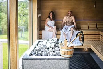 Paar sitzt in der Sauna  und im Vordergrund steht der Aufgusseimer auf dem Ofen