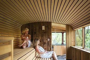 Zwei Frauen sitzen in der Schienenbus-Sauna mit dem Blick ins Freie. Im Vordergrund ist der Saunaofen