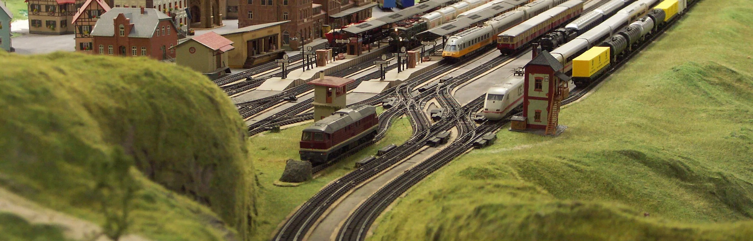Eine Miniaturlandschaft umgeben von grün. Eisenbahnschienen und ein Dorf.