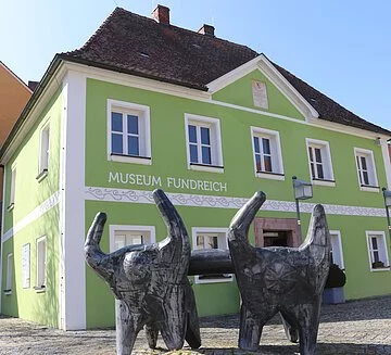 Archäologisches Museum Thalmässing Außenansicht des Archäologischen Museums in Thalmässing. Grünes Haus mit schwarzem Dach. Davor steht eine Skulptur von menschlichen Figuren, die eine Platte tragen.