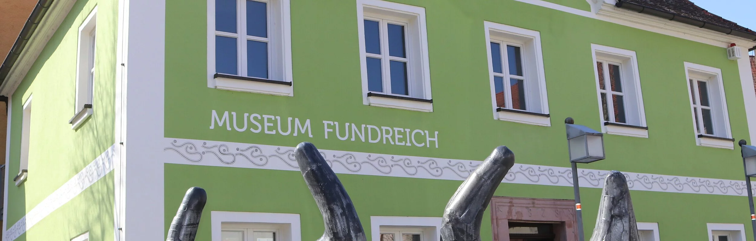Außenansicht des Archäologischen Museums in Thalmässing. Grünes Haus mit schwarzem Dach. Davor steht eine Skulptur von menschlichen Figuren, die eine Platte tragen.
