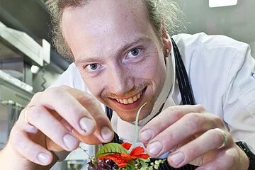 Ein junger Koch mit schwarzer Schürze und weißem Hemd lächelt in die Kamera. er dekoriert ein Tatar mit grünem Salat und einer Blume.