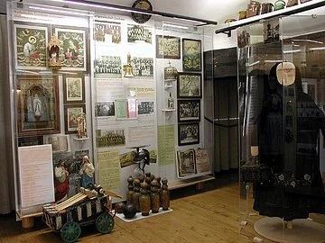 Museum Schloss Cronheim Eine Museumsausstellung mit einer alten schwarzen Tracht. Links daneben hängen christliche Bilder und beschreibende Texte an den Wänden.