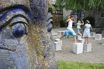 Rechts im Bild die Nahaufnahme des Gesichts einer blauen Statue. Dahinter stehen Steinwürfel in einem Hof. Drei junge Mädchen hüpfen von einem Würfel zum anderen.