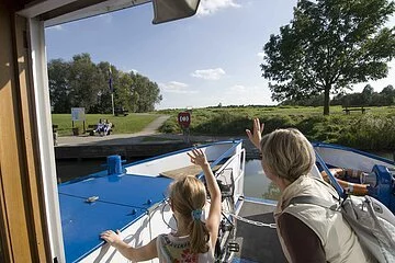 Eine Frau und ein junges Mädchen winken vom Schiff den Besuchern am grünen Uferrand.