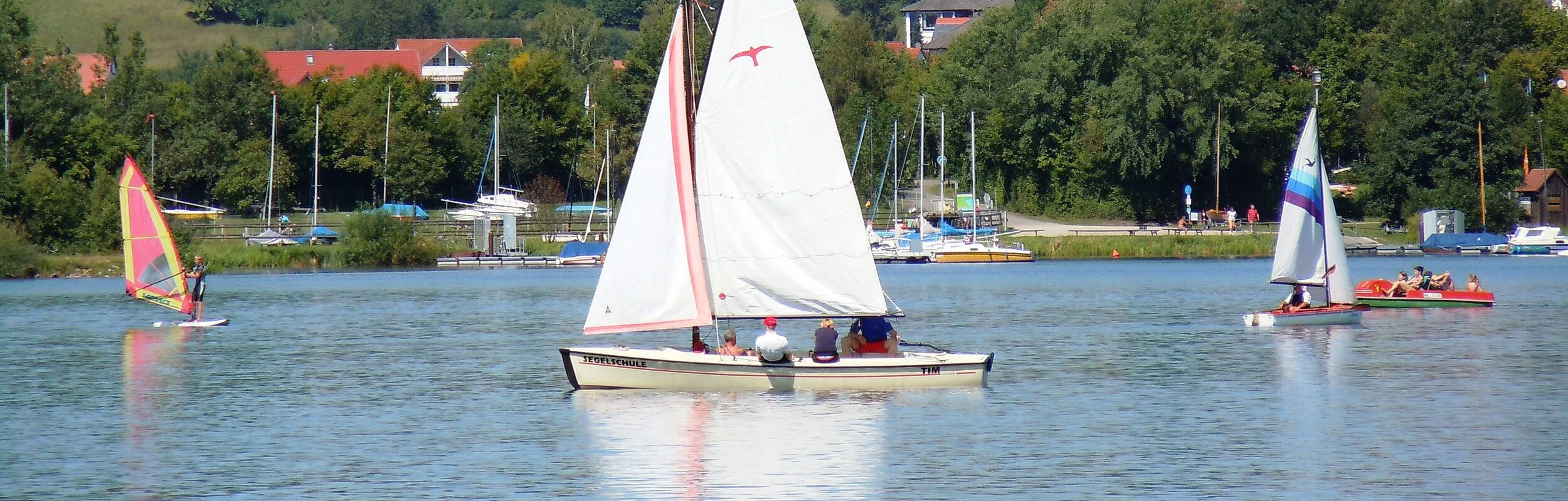 Ein weißes Segelschiff auf einem See. Daneben Windsurfer. Im Hintergund am Ufer Bäume. Etwas weiter hinten Wohnhäuser.
