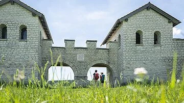 Römerkastell Vetoniana bei Pfünz Zwei Wanderer stehen am Nordtor des Römerkastells. Das Kastell wurde rekonstruiert und mit hellen Steinen gebaut. Rechts und links sind zwei Türme mit Dächern, die mit zwei Rundbögen miteinander verbunden sind.