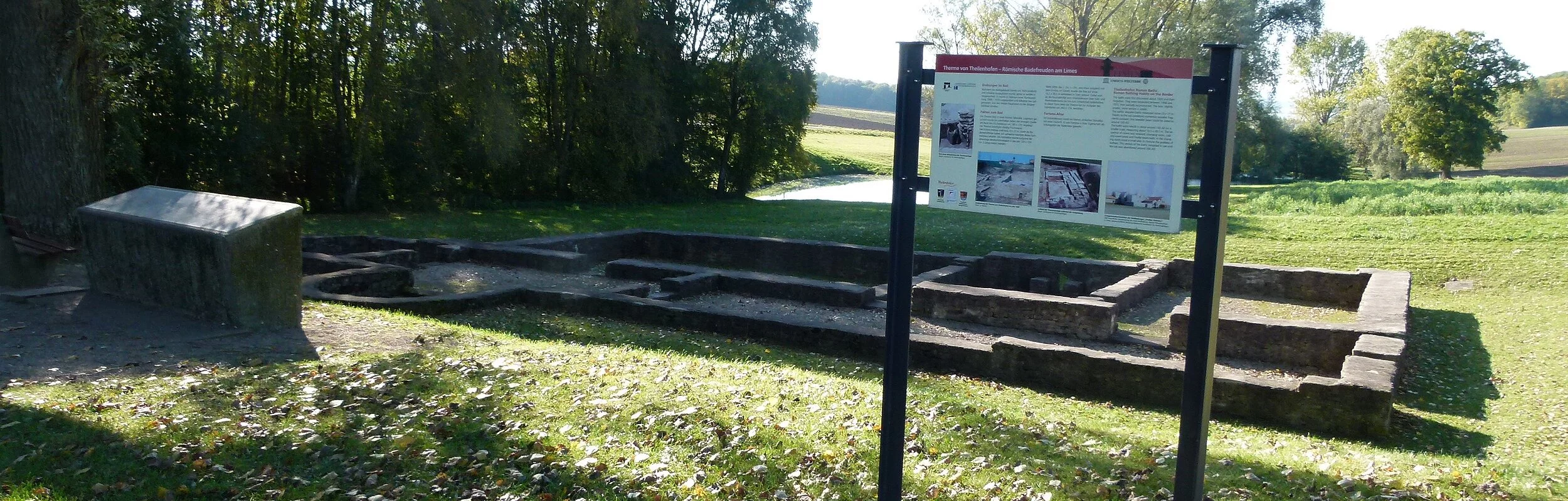 Eine römische Ruine auf einer grünen Wiese. Davor steht eine Infotafel.