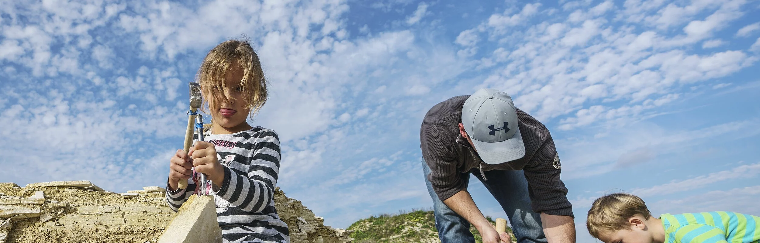 Ein Mann mit zwei Kindern suchen im Steinbruch nach Fossilien. Das Mädchen löst vorsichtig Gesteinsplatten mit Hammer und Meißel. Der Junge mit dem Mann schauen sich ihre Funde an.
