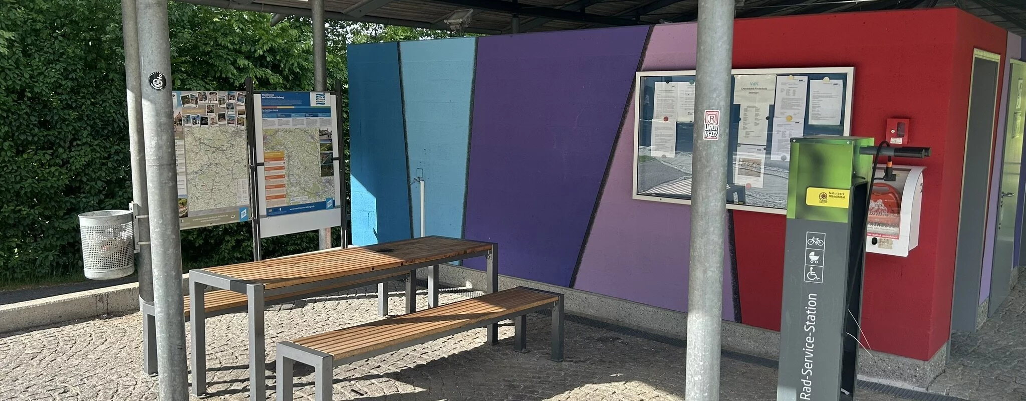 Unter dem Dach am Großparkplatz steht eine Radreparaturstation mit der man die nötigsten Reparaturen an seinem Fahrrad vornehmen kann. Links ist ein Tisch und zwei Bänke für eine kleine Rast.