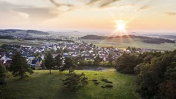 Der Blick von oben über grüne Wiesen hinab auf die Stadt Monheim. Die Sonne strahlt am Horizont und ist fast untergegangen.