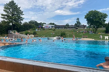Im Freibad tummeln sich Erwachsene, Kinder im Schwimmbecken. Das Wasser schimmert hellblau.