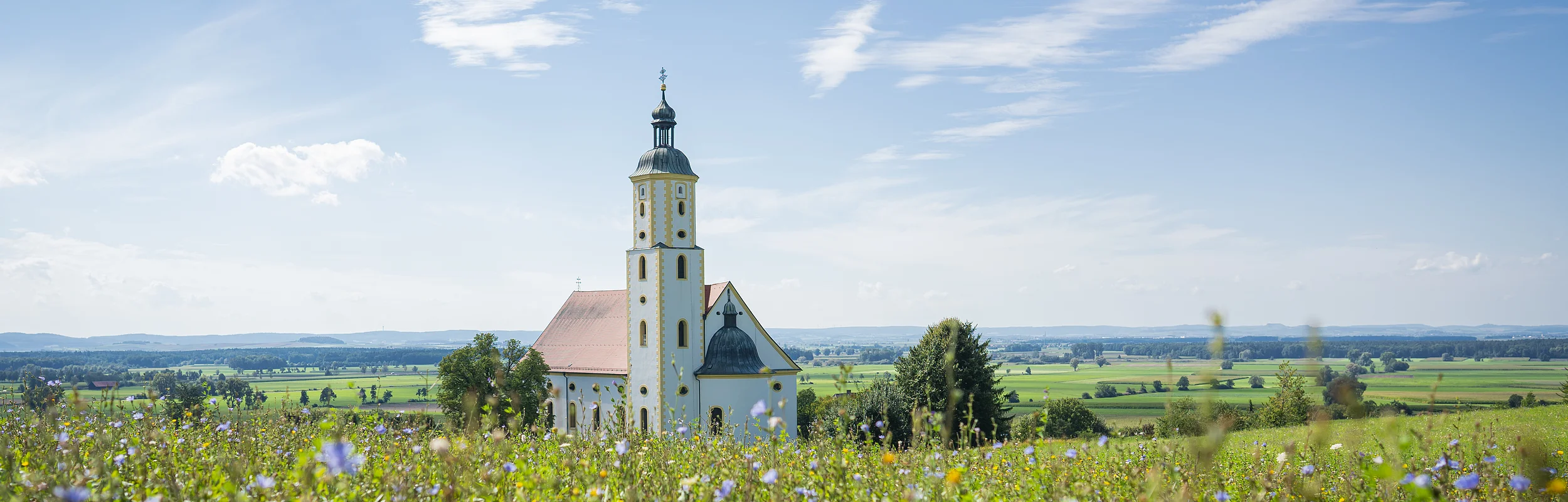 Die Wallfahrtskirche Maria Brünnlein ist von weiten zu sehen. Sie liegt idyllisch inmitten grüner Wiesen. Der Himmel strahlt blau, nur wenige Wolken sind zu sehen.