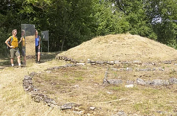Grabhügelanlage Thalmässing An der Grabhügelanlage stehen zwei Wanderer in sommerlicher Kleidung an einer Stele und bestaunen die Hügelanlage. Hinter ihnen dichter dunkelgrüner Wald.