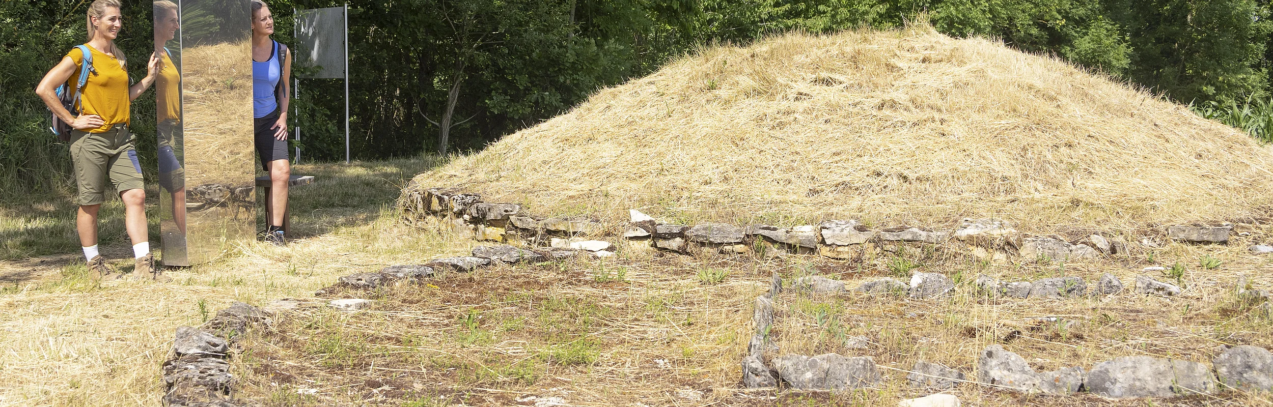 An der Grabhügelanlage stehen zwei Wanderer in sommerlicher Kleidung an einer Stele und bestaunen die Hügelanlage. Hinter ihnen dichter dunkelgrüner Wald.