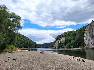 Baden an der Donau Der Kiesstrand an der Donau ist mit vereinzelten Sonnenanbetern belegt. Rechts fließt die Donau und dahinter ragen die Felswände auf. Der Himmel wäre strahlend blau, wenn nicht die vielen Wolken wären.