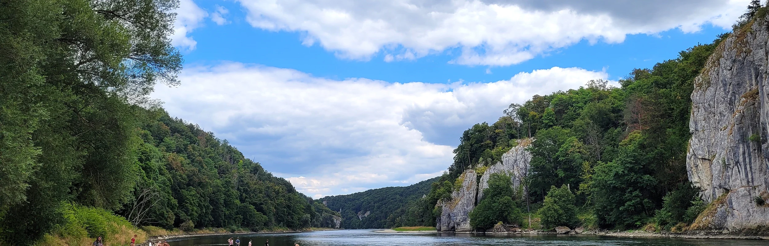 Der Kiesstrand an der Donau ist mit vereinzelten Sonnenanbetern belegt. Rechts fließt die Donau und dahinter ragen die Felswände auf. Der Himmel wäre strahlend blau, wenn nicht die vielen Wolken wären.