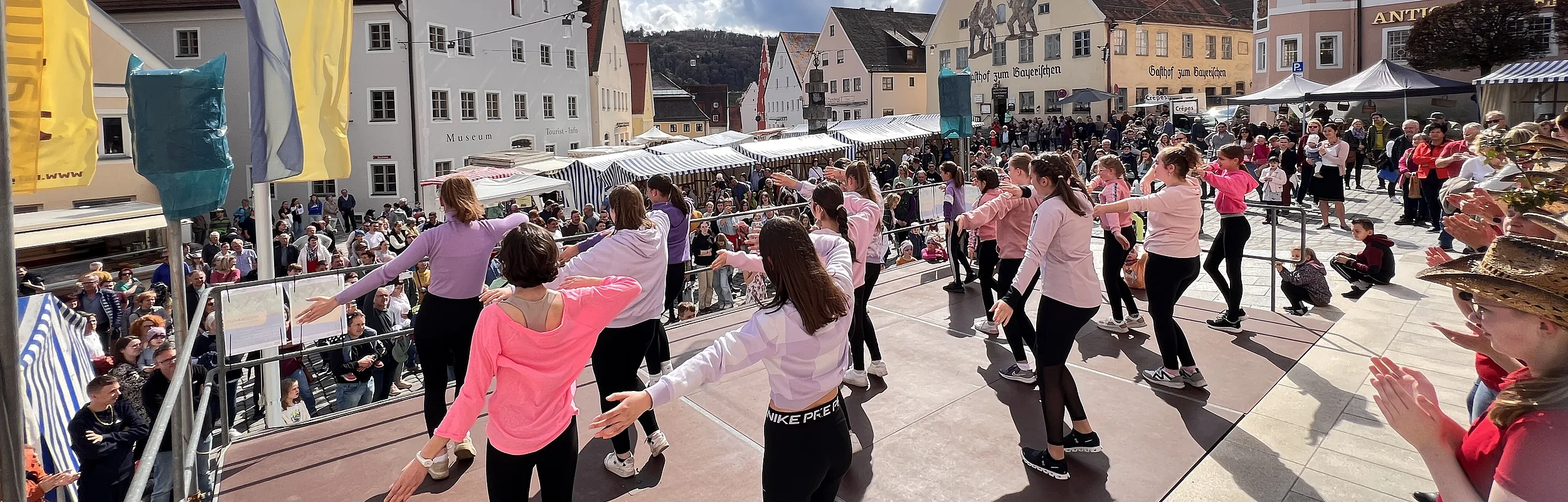 Tanzende Personen tanzen auf einer Bühne. Im Hintergrund die Altstadtkulisse von Greding, die den Rahmen des Marktplatzes bildet auf dem Marktstände und viele Besucher zu sehen sind.