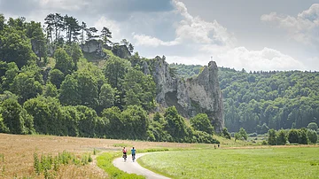 Reiseangebot Altmühltal Radweg 2025 - Standard Zwei Radfahren auf dem Altmühltal-Radweg mit Blick auf den Felsen in Dollnstein. Links und rechts sind Felder und Wiesen und die Sonne strahlt vom Himmel.