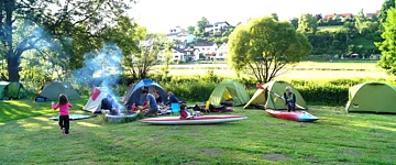 AKTIV MÜHLE GmbH & Co. KG Auf einer Wiese sind Zelte aufgebaut und in der Mitte brennt in einer Feuerschalle ein Feuer. Kinder rennen über den Platz.