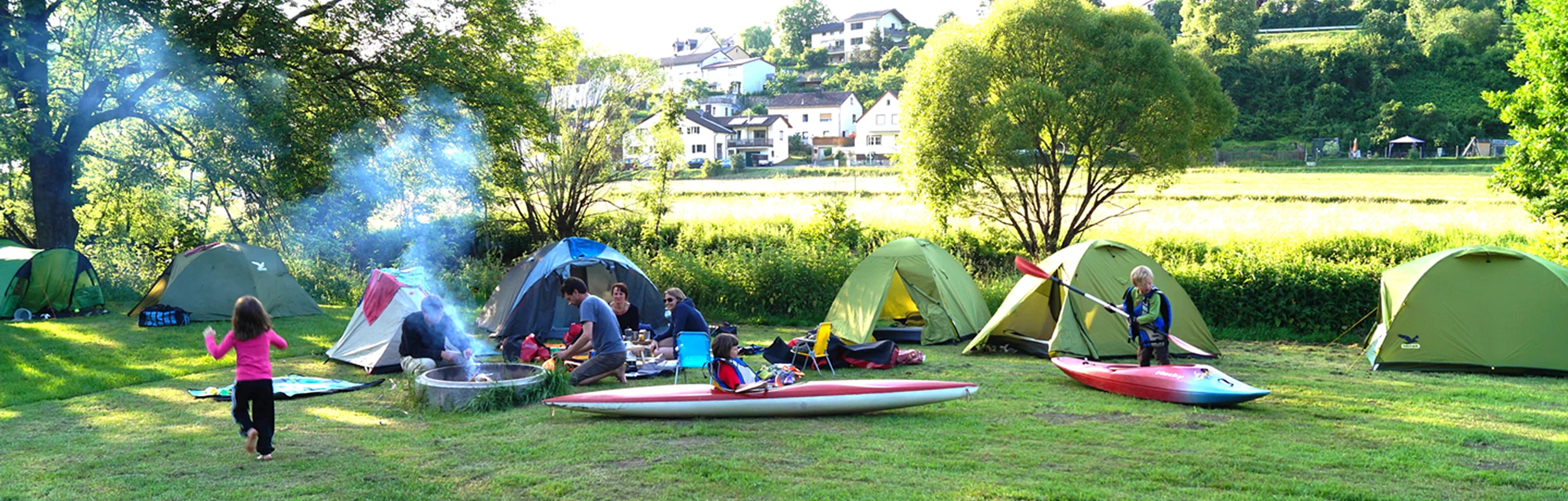 Auf einer Wiese sind Zelte aufgebaut und in der Mitte brennt in einer Feuerschalle ein Feuer. Kinder rennen über den Platz.