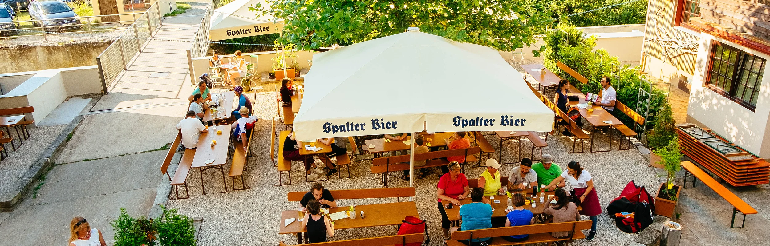 Menschen sitzen unter Sonnenschirmen auf Biergartengarnituren. Im Vordergrund parken Fahrräder.