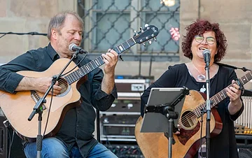 Wulli & Sonja Ein dunkel gekleideter Herr mit Gitarre in der Hand und eine dunkel gekleidete Frau mit Brille, ebenfalls mit Gitarre. Vor beiden steht ein Notenständer und zwei Mikrofone. Im Hintergrund sieht man einen Verstärker stehen und ein vergittertes Fenster.