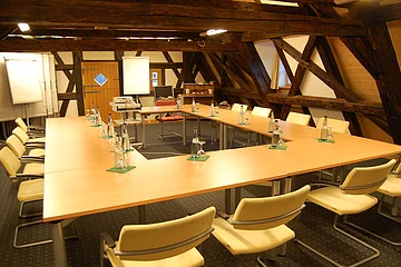 Die Holztische sind in U-Form aufgestellt. Der Raum hat viele ebenfalls holzige Deckenbalken.