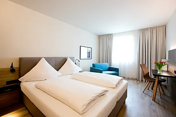 Mitten im Raum steht ein großes helles Bett, außerdem ein blauer Sessel und ein kleiner Tisch an der Wand.