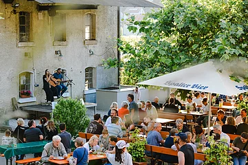 In dem Biergarten mit Bierbänke, Schirme und einer kleinen Bühne vor einem Haus der Aktiv Mühle sitzen viele Menschen gemütlich auf Ihren Plätz und hören der Musik auf der Bühne zu.