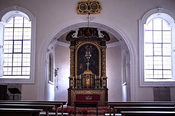 Der prunkvolle Altar steht inmitten eines Gewölbebogens, vor ihm stehen einige Bänke.