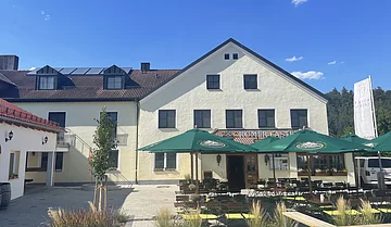 Vor dem hellen Gebäude befindet sich ein Biergarten mit grünen Sonnenschirmen.
