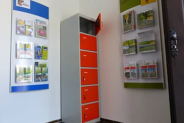 Ein orange-grauer Indoor-Ladeschrank für eBike-Akkus mit insgesamt sechs Fächern und einem offenen Fach steht zwischen zwei Wanddisplays mit Broschüren in dem Vorraum der Touristinformation Monheim.