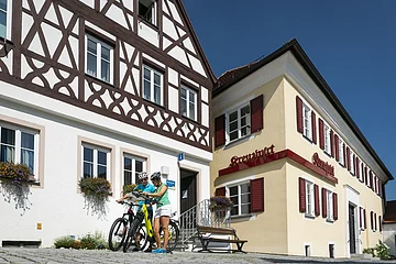 Tourist-Information der Stadt Monheim/Monheimer Alb: ein Fachwerkhaus mit drei Stockwerken, weißen Wänden und braunen Balken. Rechts davon ein gelbes zweistöckiges Gebäude mit roten Fensterläden. Zwei Personen mit Fahrrädern stehen davor und schauen eine Broschüre an.