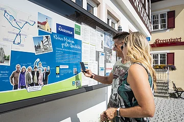 Zwei Personen vor einem weißen Gebäude, der Touristinformation Monheim betrachten eine Informationstafel mit einer Karte und Texten zur Lauschtour in Monheim und haben ein Smartphone in der Hand, mit dem sie einen Code abscannen.