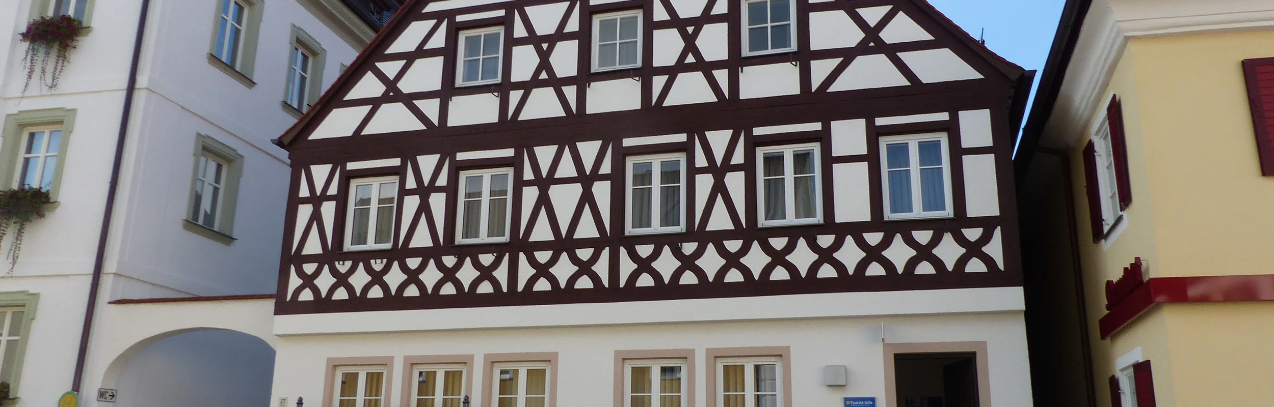 Tourist-Information der Stadt Monheim/Monheimer Alb: ein Fachwerkhaus mit drei Stockwerken, weißen Wänden und braunen Balken. Davor steht eine Sitzbank und ein Schaukasten auf einem gepflasterten Platz. Links davon ein weißes dreistöckiges Gebäude. Rechts davon ein gelbes zweistöckiges Gebäude mit roten Fensterläden.