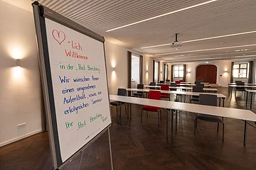 Post Berching Veranstaltungsraum Die Tafel steht vorne, im Raum sind verschiedenfarbige Stühle an weißen Tischen verteilt.