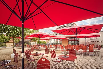Biergarten Post Berching Unter den roten Sonnenschirmen stehen Holztische mit Holzstühlen.