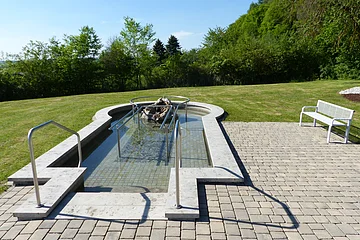Ein befülltes Kneippbecken mit Handlauf, umgeben von Rasen und Bäumen, daneben eine weiße Bank auf Pflastersteinen.
